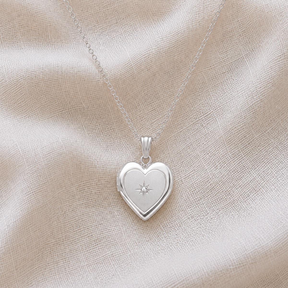 A sterling silver heart-shaped locket pet memorial necklace with a small starburst with diamond detail in the center, resting on a textured beige fabric background.