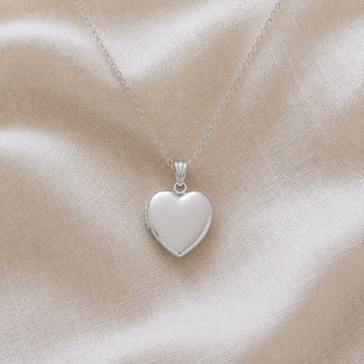 A polished sterling silver heart-shaped pendant locket pet memorial necklace on a delicate silver chain, displayed on a draped beige fabric background.