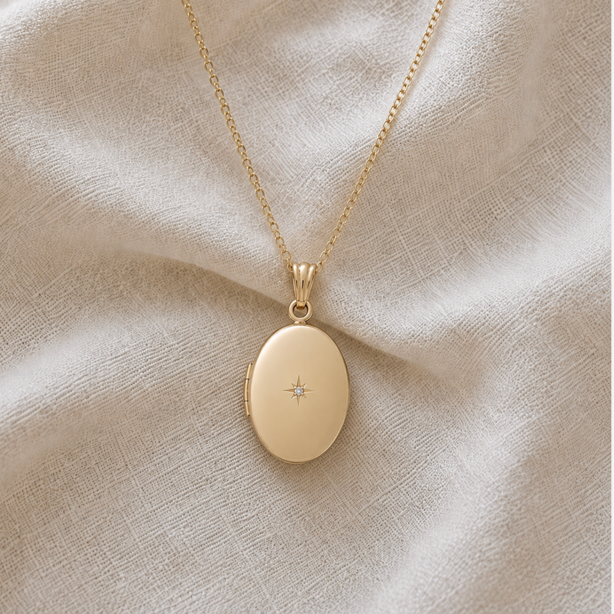 A 24K gold filled oval locket pendant necklace with a tiny starburst with diamond in the center, resting on a textured beige fabric background.