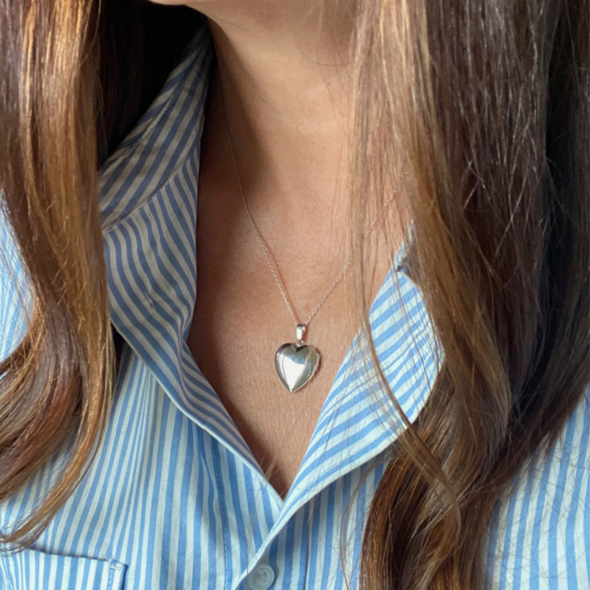 A close-up of a person wearing a sterling silver heart pet memorial locket necklace wearing a light blue and white striped button-down shirt.