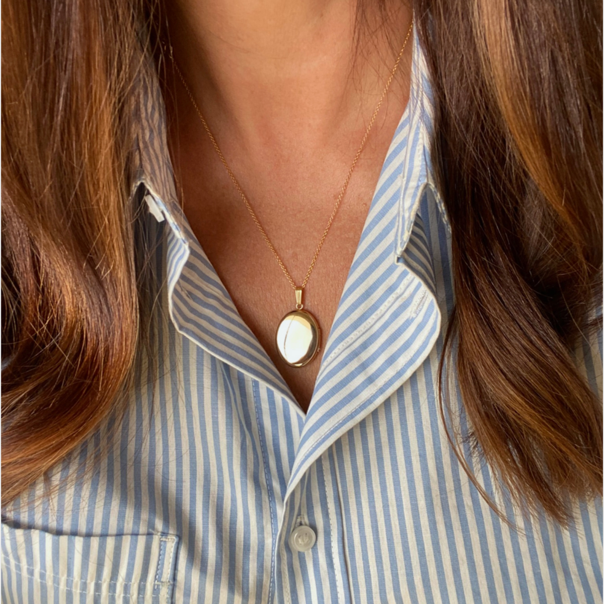 A close-up of a woman wearing a gold oval locket necklace, styled with a blue and white striped button-down shirt.