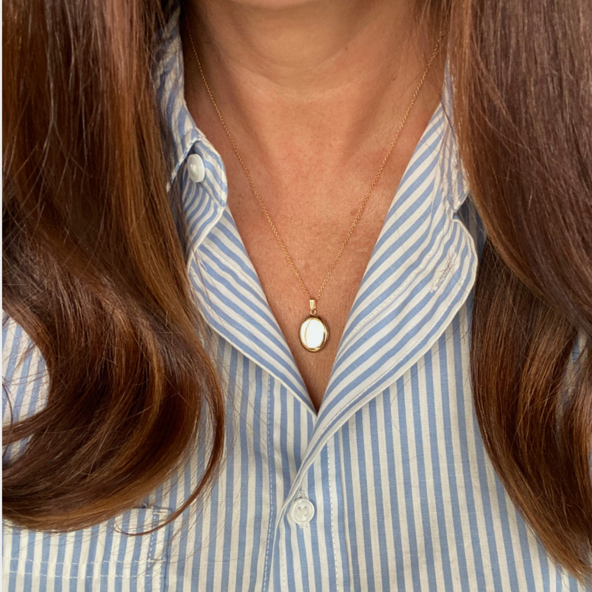 A close-up of a person wearing a Petite 14K gold-filled oval pet memorial locket on a delicate gold chain, styled with a blue and white striped button-down shirt.