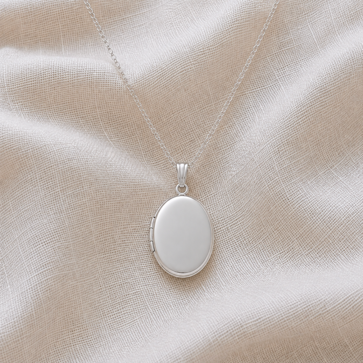 A sterling silver oval locket pet memorial necklace with a smooth, blank center, resting on a textured beige linen fabric background.