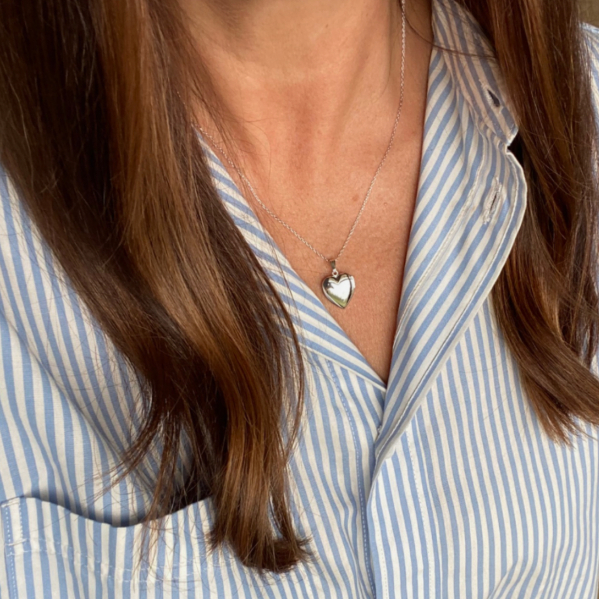 A close-up of a woman wearing a sterling silver heart locket pet memorial necklace over a light blue and white striped button-down shirt.