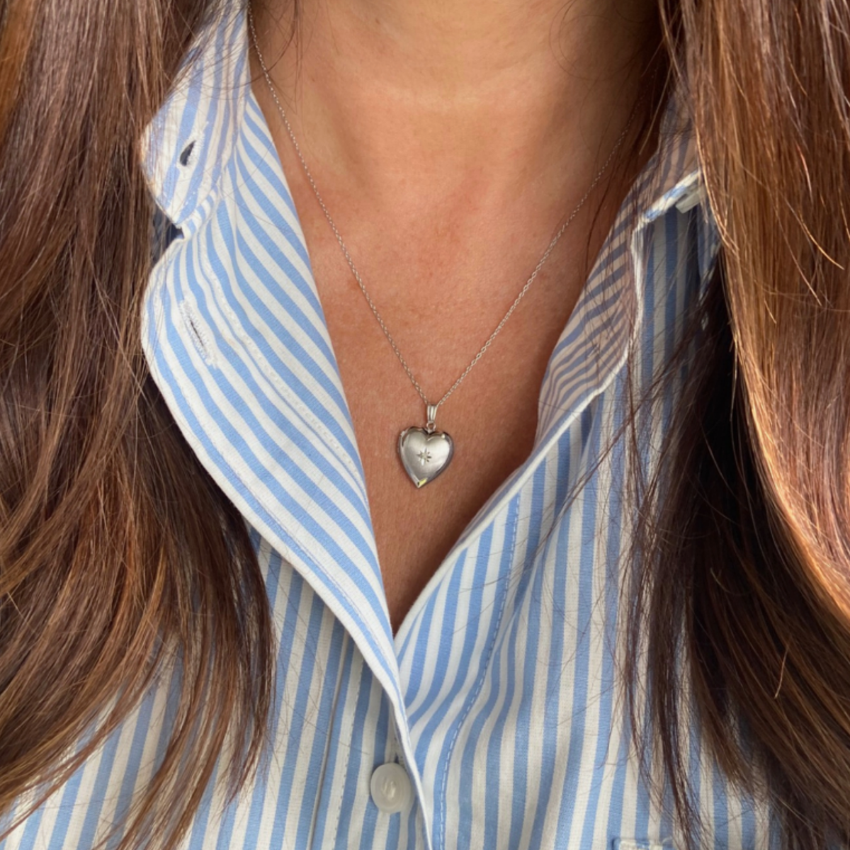 A close-up of a person wearing a sterling silver heart locket necklace with a diamond in the center, over a light blue and white striped button-down shirt.