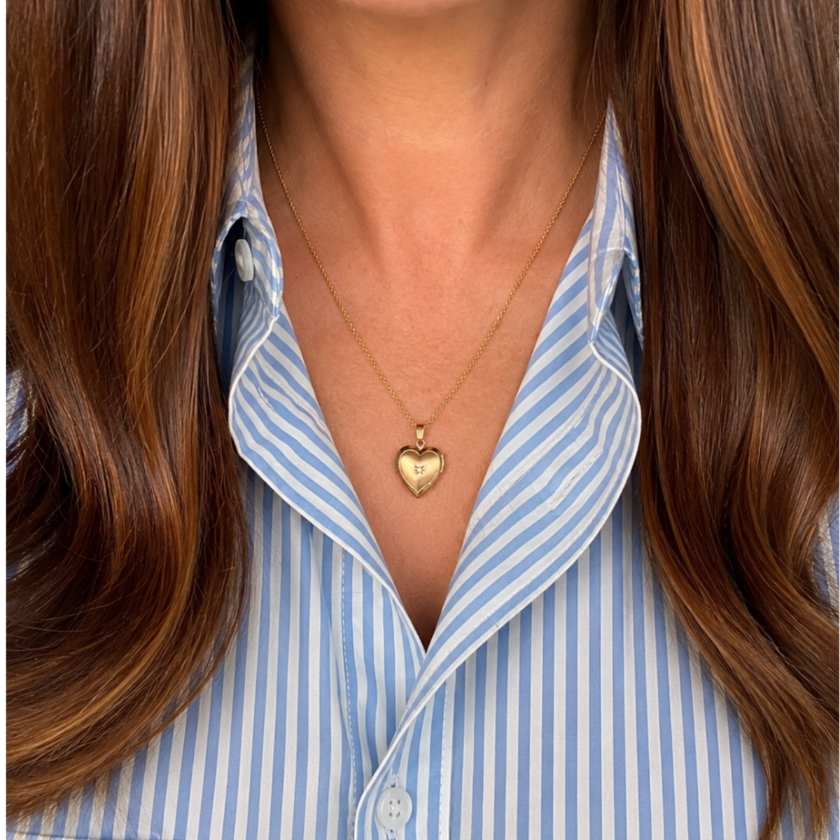 A close-up of a person wearing a delicate 14k gold filled heart locket necklace over a light blue and white striped button-down shirt.