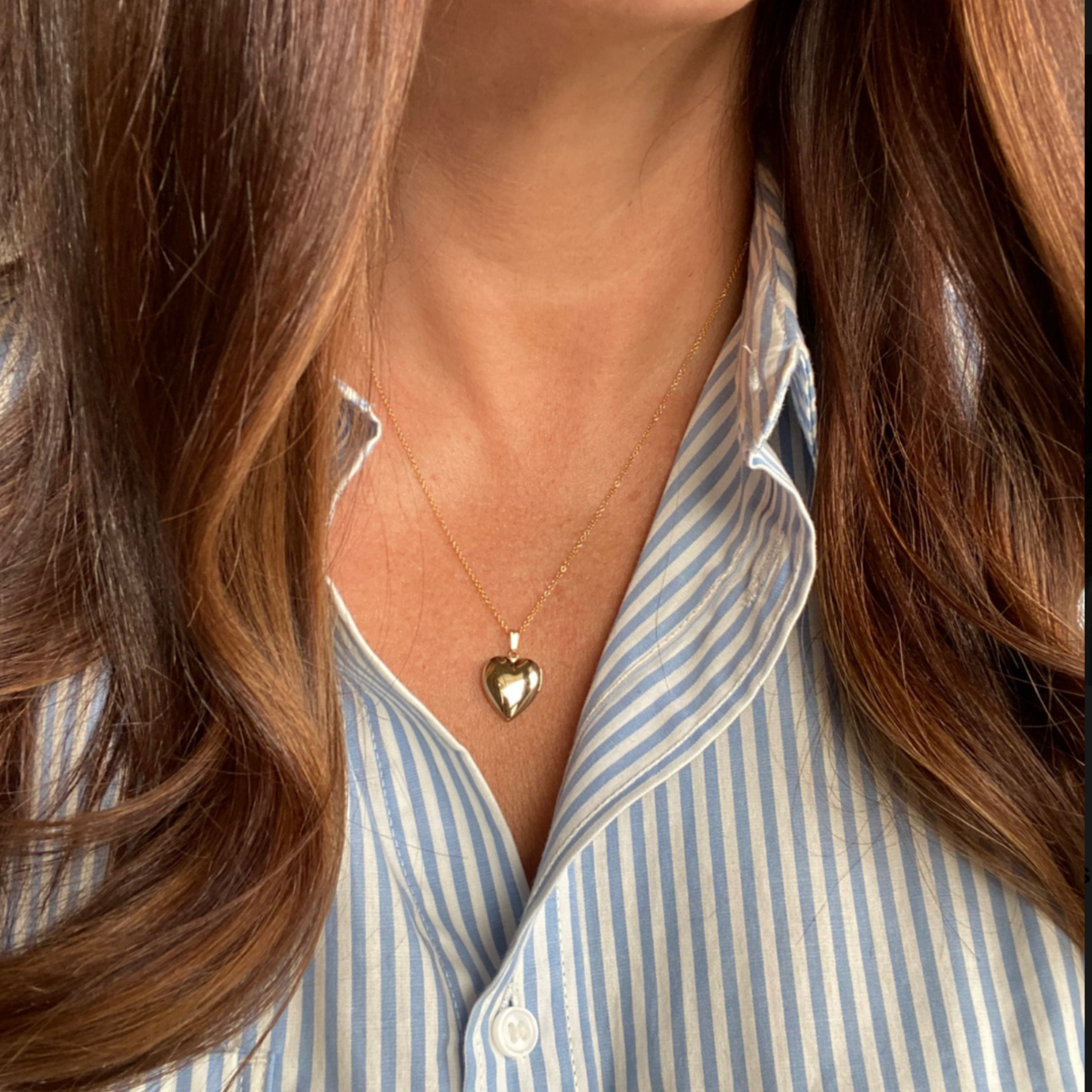 A close-up of a person wearing a delicate 14k gold filled gold heart locket  necklace over a light blue and white striped button-down shirt.