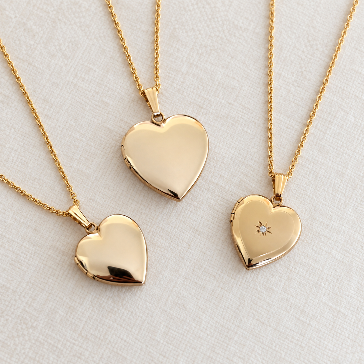 Three gold heart-shaped locket necklaces on delicate chains, displayed on a textured off-white fabric background. One necklace features a polished heart pendant, another has a heart with a tiny clear stone, and the third is shown open to reveal a heart-shaped compartment.