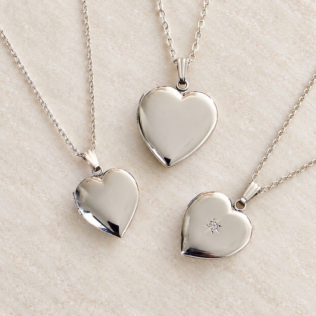 Three silver heart-shaped locket necklaces with chains, displayed on a light beige textured surface. One pendant is plain, one has a small star detail, and one is shown closed.
