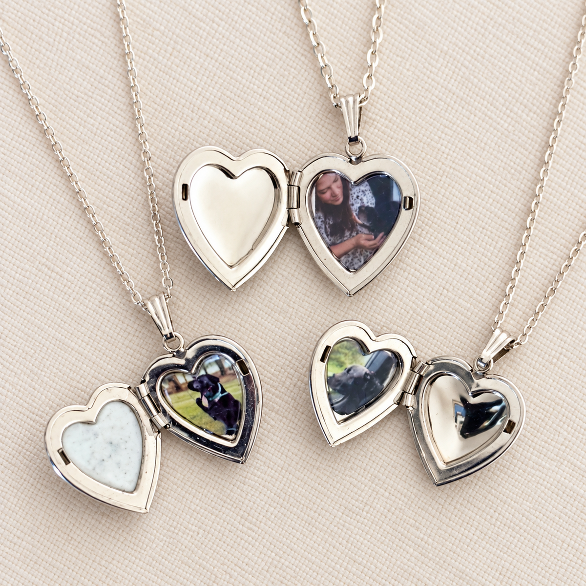 Four silver heart-shaped locket necklaces on chains, arranged on a light beige fabric background. Three are open to show custom photo inserts, and one is closed showing a light blue stone insert.