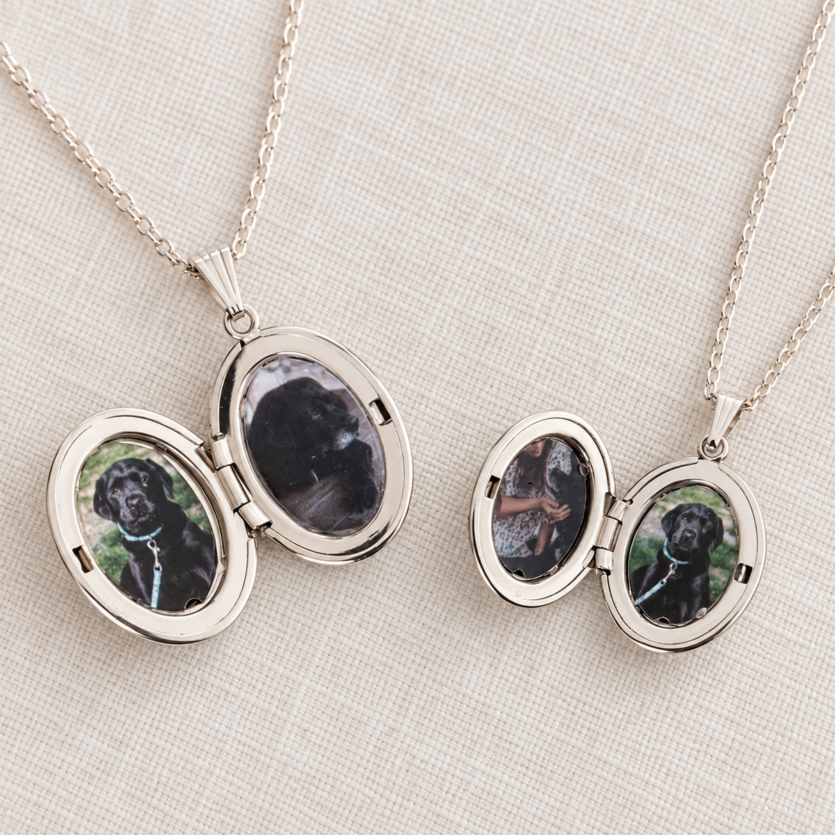 Two silver oval locket necklaces on a light beige fabric background, each open to show a custom photo insert of a black dog.